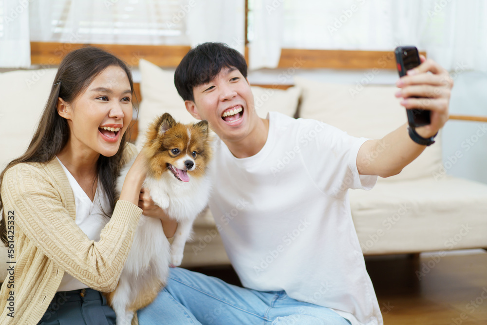 Happy cheerful Asian young couple taking a selfie photography or making a selfie video call on smartphone with their little dog in living room. 