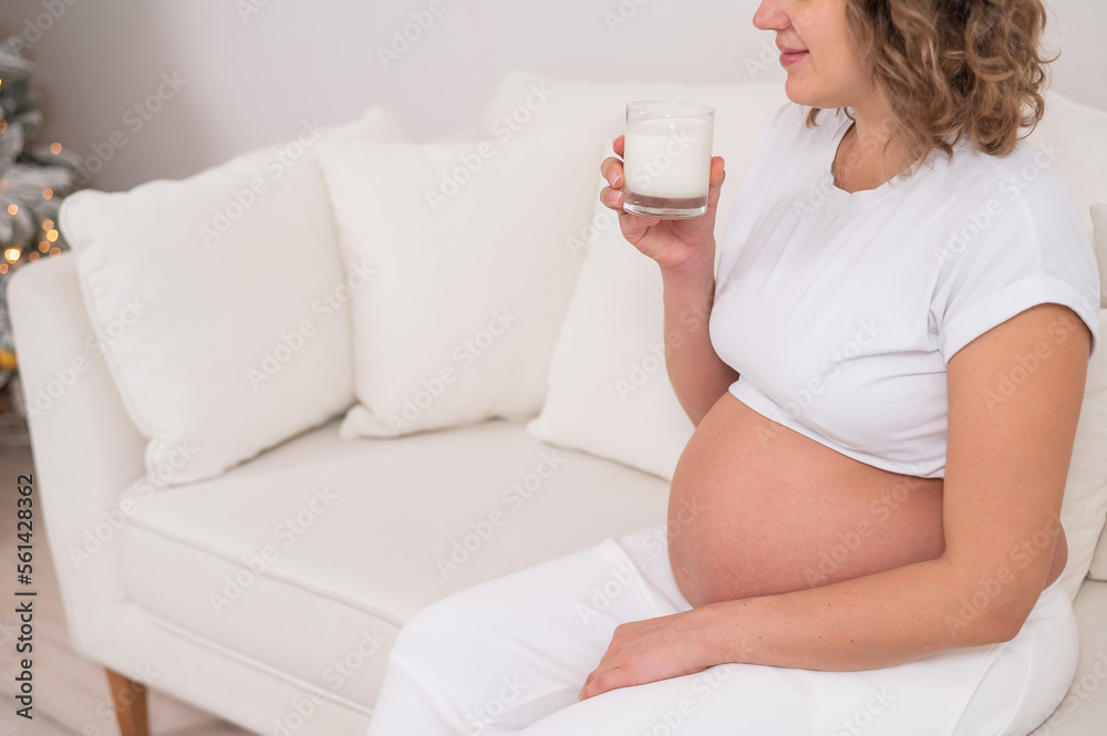 A pregnant woman sits on a white sofa and drinks milk.