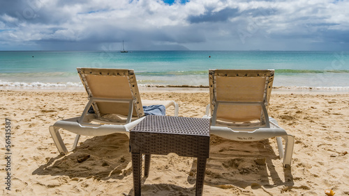 Two sun loungers and a wicker table on a tropical beach. Footprints in the sand. Ahead is the turquoise ocean. Silhouettes of yachts and islands on the horizon. Clouds in the blue sky. Seychelles.