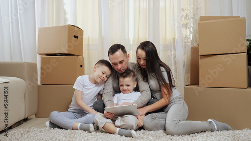 Excited family sits on floor among boxes and looks at tablet. Parents and boy children enjoy spending together weekend in new apartment living room