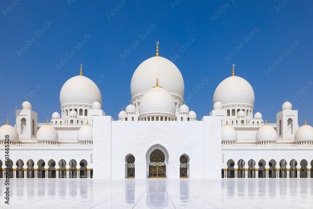 Symmetrical close up of the main courtyard of the Sheik Zayed grand mosque in Abu Dhabi with the prayer hall and its round domes against a blue sky