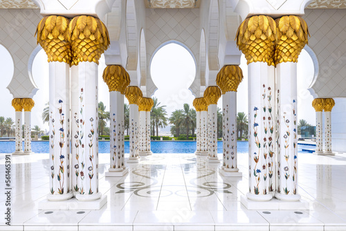 A white marbe colonnade with golden capitals leading to the external pool with palm trees on the background at Sheik Zayed mosque