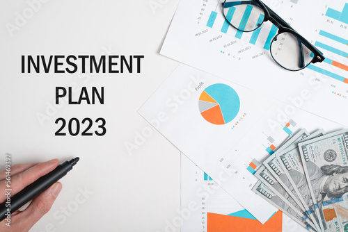 Investment, business and finance concept. Close-up of hand writing Investment Plan 2023 with black marker, papers with graphs, money and glasses at workplace
