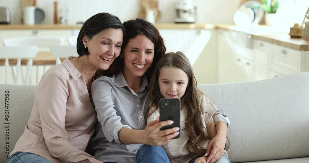 Multi generational women, happy family rest on sofa enjoy virtual meeting, talk to friend or relatives living abroad use videocall, staring at cellphone screen laughing have fun use mobile application Multi generational women, happy family rest on sofa enjoy virtual meeting, talk to friend or relatives living abroad use videocall, staring at cellphone screen laughing have fun use mobile application