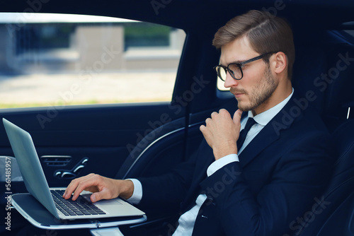 Thoughtful young businessman keeping hand on chin while sitting in the lux car and using his laptop. © ty