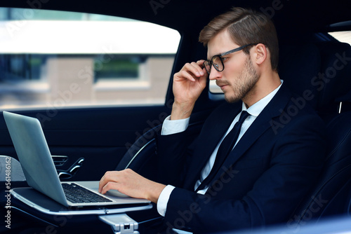Thoughtful confident businessman keeping hand on glasses while sitting in the luxe car and using his laptop. © ty