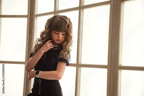 Sad image unhappy little girl in stylish black dress posing near large window indoors, closed eyes. Studio shot pensive teen cute lady. Child emotion concept. Copy text space for advertising
