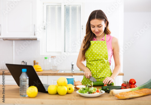 Positive woman cooking dinner and using laptop