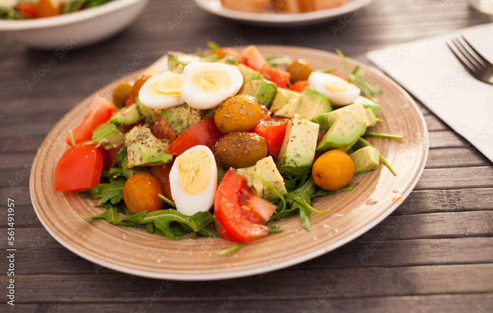 fresh salad of arugula, avocado, cherry tomatoes with olives and quail eggs on brown plate