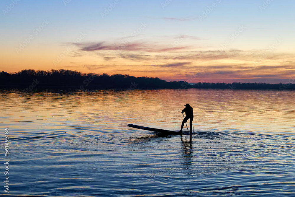 Silhouette of young athlete standing on SUP (stand up paddle board) at autumn sunset in the Danube river. Water sport at blue hour