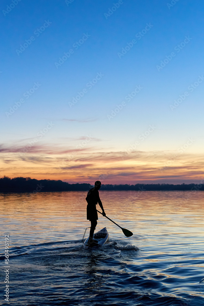 Fototapeta premium Silhouette of young man standing on SUP (stand up paddle board) at autumn sunset in the Danube river. Water sport at blue hour