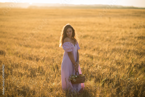 a girl with long dark hair in a lilac dress with a basket in her hands at sunset in a wheat field