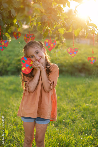 A cute girl is standing in the park at sunset holding a postcard with colored puzzles in her hand. A tree decorated with hearts with jigsaw pieces. Autism Awareness Day 