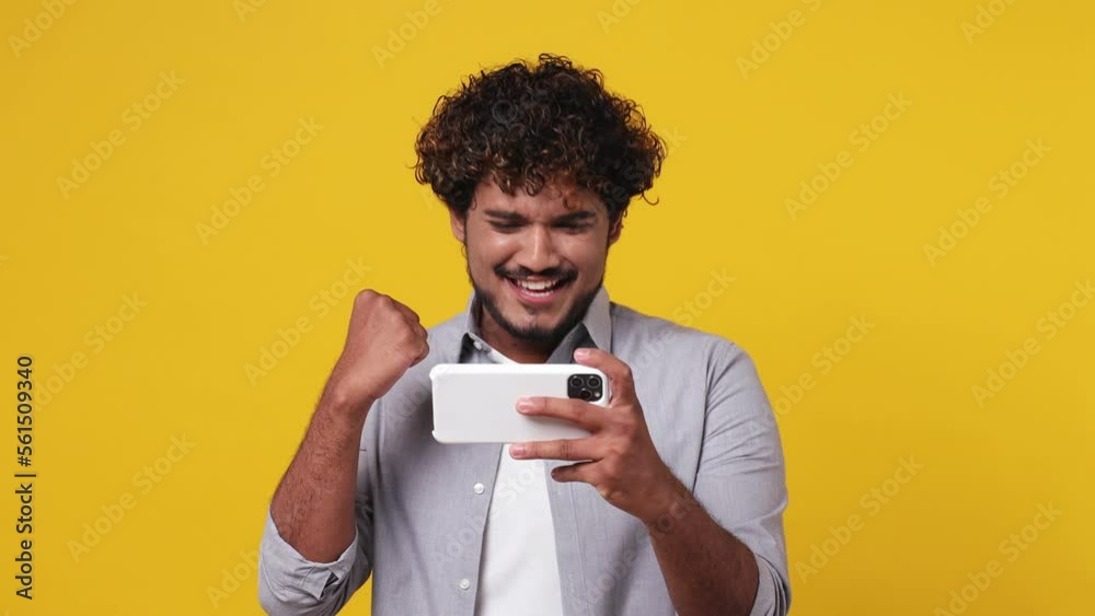 Gambling excitable young Indian man in casual grey shirt use play racing app on mobile cell phone hold gadget smartphone for pc video games do winer gesture isolated on plain yellow background studio