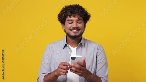 Dreamful pensive minded young Indian man 20s wear casual grey shirt hold use mobile cell phone typing browsing chatting send sms isolated on plain yellow background studio. People lifestyle concept photo