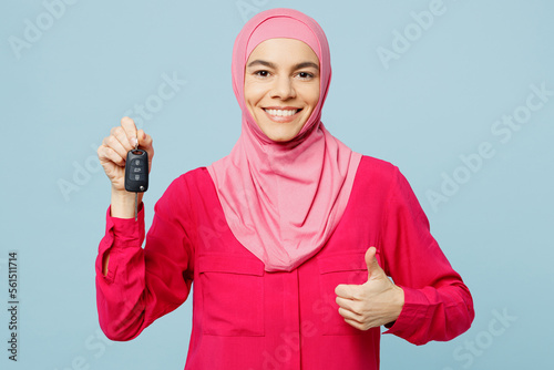 Young smilng arabian muslim woman wears pink abaya hijab hold car keys fob keyless system show thumb up isolated on plain pastel light blue cyan background studio People uae islam religious concept.