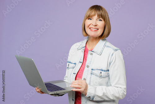 Elderly smiling cheerful blonde IT woman 50s year old she wear casual clothes denim jacket t-shirt hold use work on laptop pc computer isolated on plain pastel light purple background studio portrait