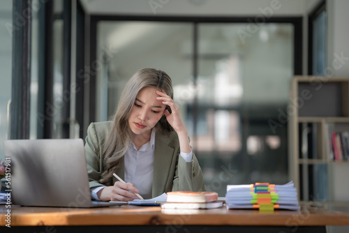 Sad Asian woman looking annoyed and stressed, sitting at the desk, using a laptop, thinking, feeling tired and bored with depression problems © Songsak C