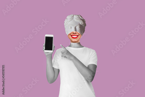 Young smiling woman headed by antique statue with red lips holds mobile phone and points at black screen demonstrating copy space for text or design isolated on purple background. Contemporary art photo