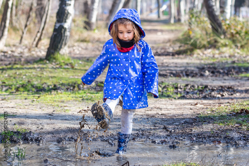 Little caucasian girl playing with the puddles created by the rain. Girl in a raincoat and rain boots. Children's games in the park.