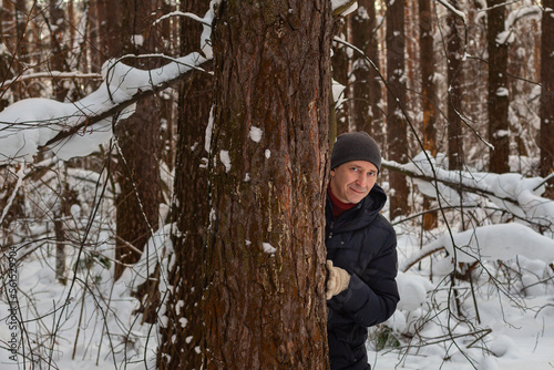 Portrait of young millennial smiling man peeking out from pine trunk in winter coniferous forest.