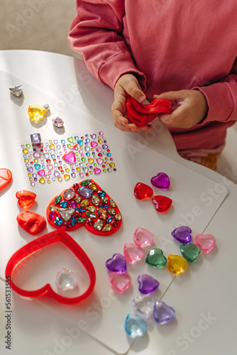 Child hands creating red heart from play dough for modeling with decorate from crystal rhinestones and shiny stones. Toddlers crafts for Valentine's Day. Holiday Art Activity for Kids. © igishevamaria