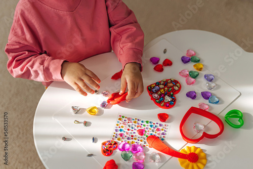 Child hands creating red heart from play dough for modeling with decorate from crystal rhinestones and shiny stones. Toddlers crafts for Valentine's Day. Holiday Art Activity for Kids.