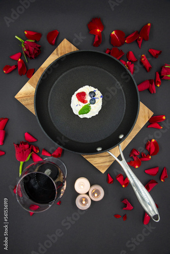 Valentine's day concept with red rose flower petals on a black background and a pan placed on a wooden board and a pavlova cake inside the pan. A glass of red wine next to it. View from above. photo