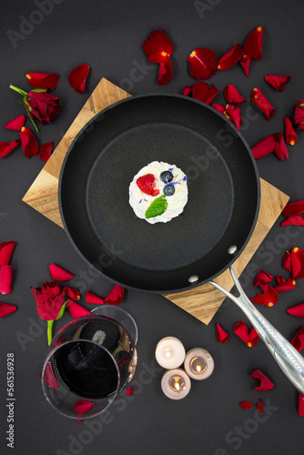 Valentine's day concept with red rose flower petals on a black background and a pan placed on a wooden board and a pavlova cake inside the pan. A glass of red wine next to it. View from above.