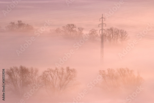 High voltage power lines emerging from ground fog in an agricultural landscape. Pastel colours of sunrise, warm tones, ground fog, frosty morning. Czech Republic.