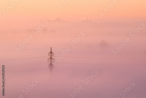 High voltage power lines emerging from ground fog in an agricultural landscape. Pastel colours of sunrise, warm tones, ground fog, frosty morning. Czech Republic.