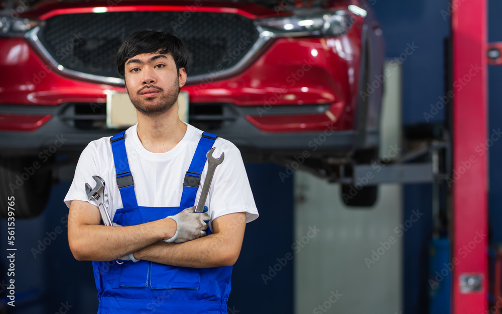 Adult Asian handsome male mechanic wearing uniform, crossed arms, posing with confidence, standing in garage at car or automobile maintenance service center or shop with copy space. Industry Concept. Adult Asian handsome male mechanic wearing uniform, crossed arms, posing with confidence, standing in garage at car or automobile maintenance service center or shop with copy space. Industry Concept.