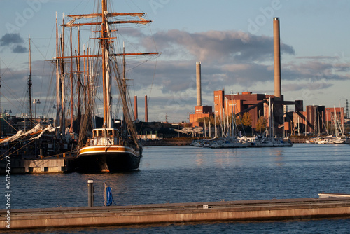 Un des ports d'Helsinki avec de nombreux voiliers, dont un ancien 3 mâts. En arrière plan, une usine avec de grandes cheminées industrielles. La scène est éclairée par la lumière d'une soleil de fin d