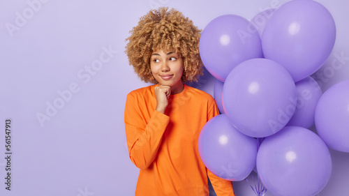 Horizontal shot of thoughtful woman dressed in orange jumper keeps hand under chin concentrated aside holds bunch of inflated balloons thinks how to celebrate birthday isolated over purple background