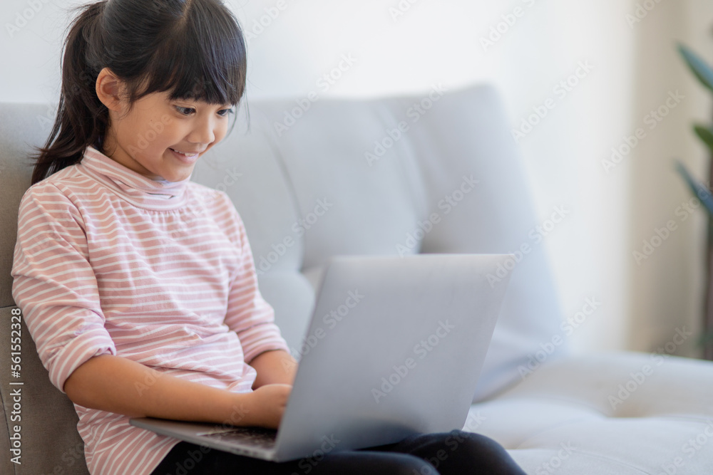 Asian Little school kid girl use laptop computer sitting on sofa alone at home. Child learning reading online social media content, play education lessons game chatting with friends. Asian Little school kid girl use laptop computer sitting on sofa alone at home. Child learning reading online social media content, play education lessons game chatting with friends.