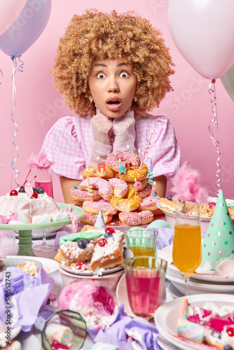 People and celebration concept. Shocked curly haired adult woman wears dress and gloves surrounded by desserts celebrates special occasion isolated over pink background finds out surprising news