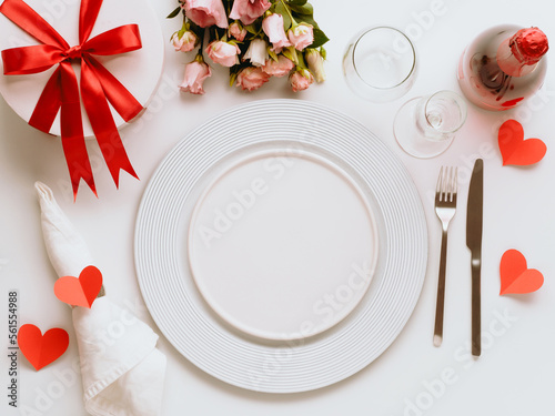 Valentines Day tablesettings concept on white background. Empty white plates, gold cutlery, rose wine bottle, glasses, flowers bouqet, gift box. Copy space. Topview flatlay