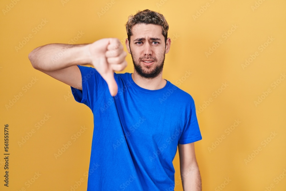 Hispanic man with beard standing over yellow background looking unhappy and angry showing rejection and negative with thumbs down gesture. bad expression.