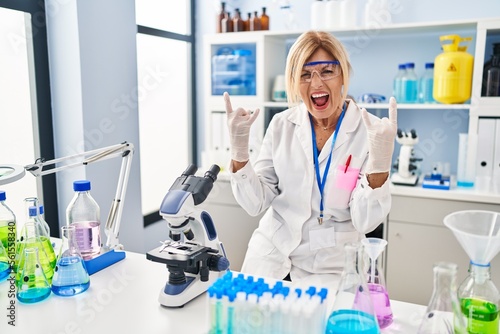 Middle age blonde woman working at scientist laboratory shouting with crazy expression doing rock symbol with hands up. music star. heavy music concept.