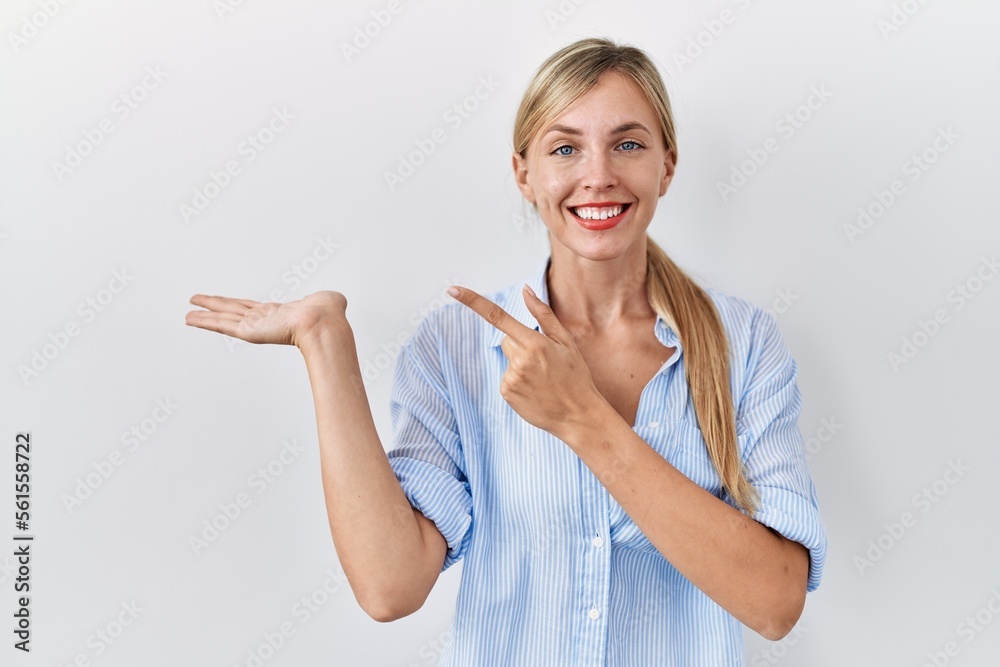 Beautiful blonde woman standing over white background amazed and smiling to the camera while presenting with hand and pointing with finger. Beautiful blonde woman standing over white background amazed and smiling to the camera while presenting with hand and pointing with finger.