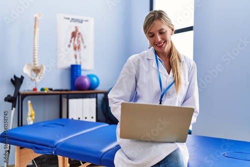 Young blonde woman wearing physiotherpist uniform using laptop at physiotherapy clinic