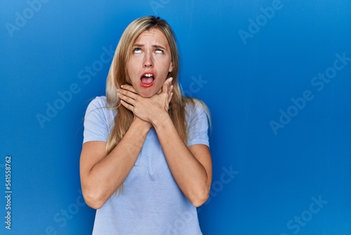 Beautiful blonde woman wearing casual t shirt over blue background shouting and suffocate because painful strangle. health problem. asphyxiate and suicide concept.