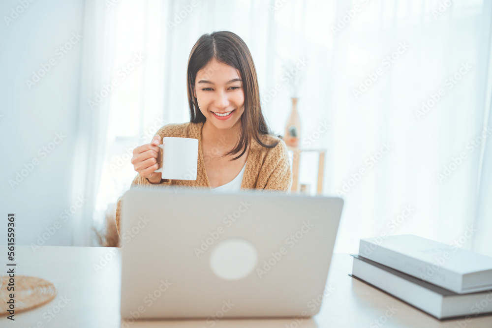 Smiling young asian woman using laptop while holding a cup of coffee at home. lifestyle Smiling young asian woman using laptop while holding a cup of coffee at home. lifestyle