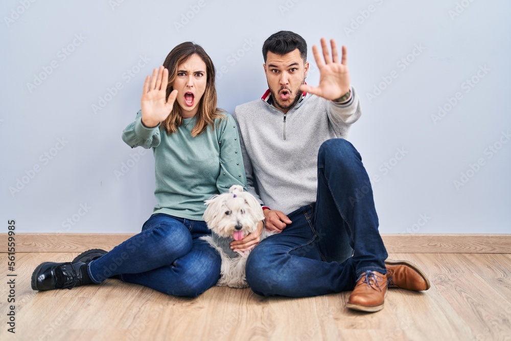 Young hispanic couple sitting on the floor with dog doing stop gesture with hands palms, angry and frustration expression Young hispanic couple sitting on the floor with dog doing stop gesture with hands palms, angry and frustration expression