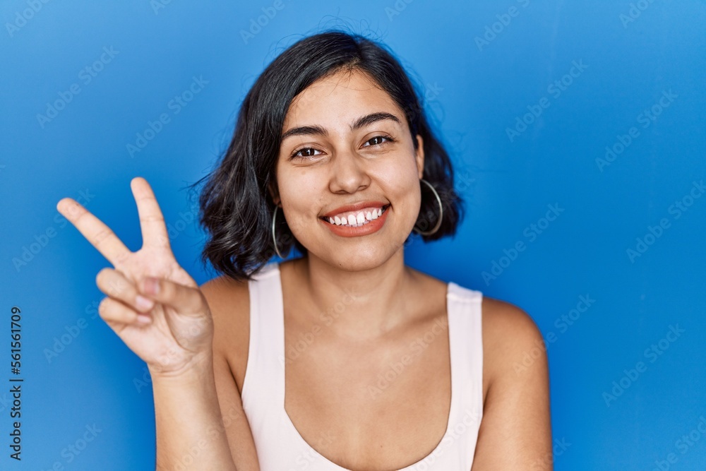 Fototapeta premium Young hispanic woman standing over blue background smiling with happy face winking at the camera doing victory sign. number two.