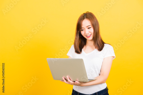 Asian happy portrait beautiful cute young woman teen smiling standing wear t-shirt using laptop computer on hand looking to computer isolated, studio shot on yellow background with copy space © sorapop