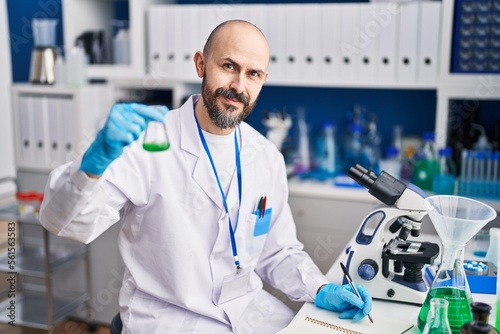 Young bald man scientist holding test tube writing on notebook at laboratory