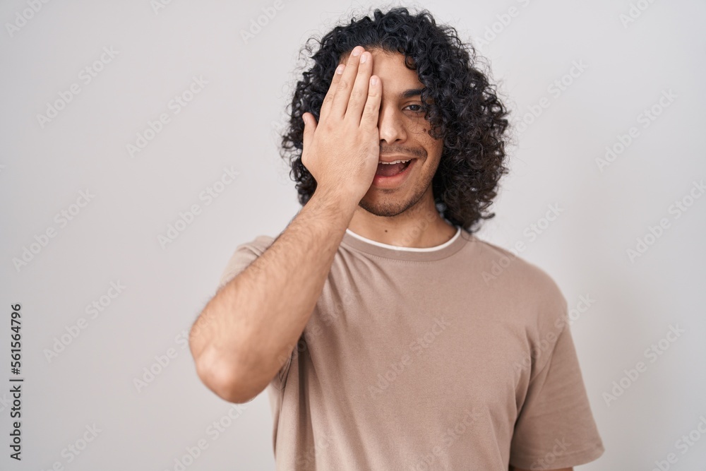 Hispanic man with curly hair standing over white background covering one eye with hand, confident smile on face and surprise emotion. Hispanic man with curly hair standing over white background covering one eye with hand, confident smile on face and surprise emotion.