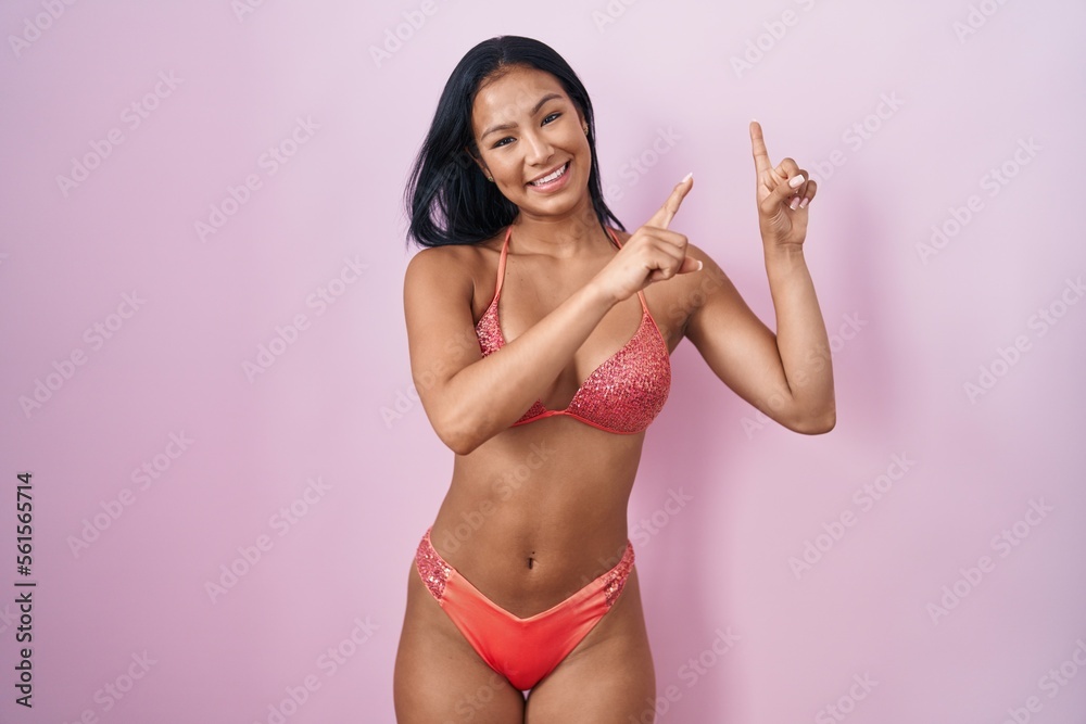 Naklejka premium Hispanic woman wearing bikini smiling and looking at the camera pointing with two hands and fingers to the side.