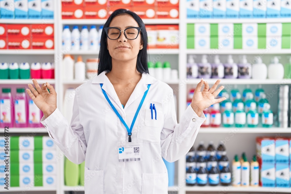 Hispanic woman working at pharmacy drugstore relax and smiling with eyes closed doing meditation gesture with fingers. yoga concept.